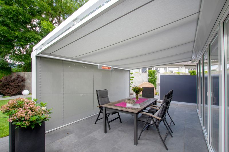 White patio roof with under-glass awning markilux 779 with white frame and gray fabric cover. In addition, a vertical blind with the same fabric pattern and an infrared heater are mounted at the front.